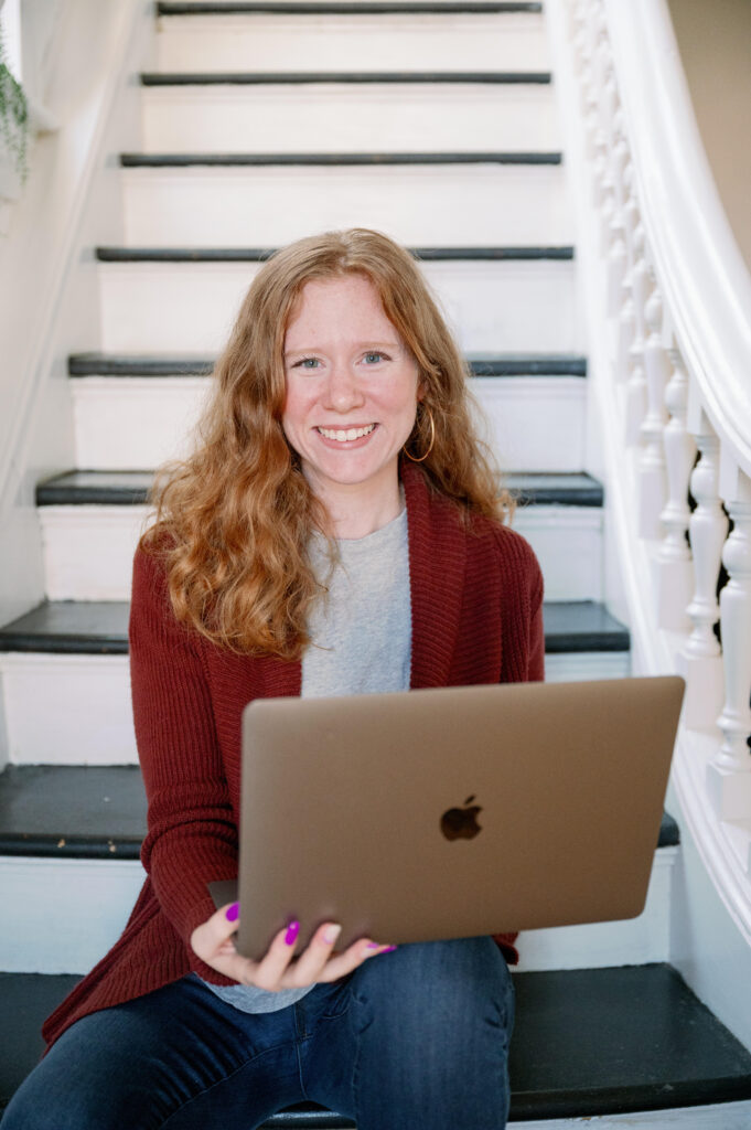 Image of Haley, she is seated on a stair case, holding a laptop and smiling into the camera